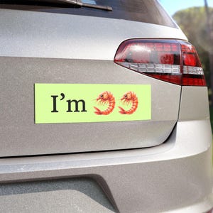 May include: A rectangular, light green car magnet with the text "I'm" in black, followed by two watercolor shrimp illustrations. The magnet is affixed to the back of a silver car.