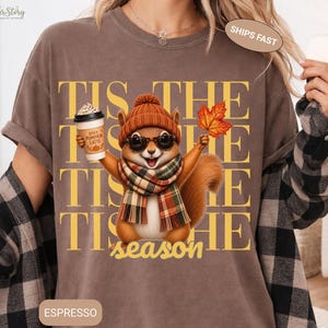 May include: A brown t-shirt featuring a graphic of a squirrel wearing an orange knit hat, sunglasses, and a plaid scarf. The squirrel holds a coffee cup labeled Spicy Pumpkin Latte and an orange maple leaf. The text reads TIS THE SEASON.