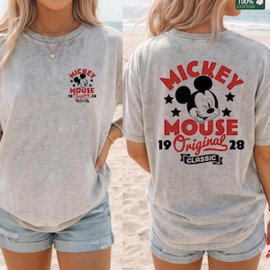 May include: A light gray t-shirt featuring a Mickey Mouse graphic on the front and back. The design includes Mickey's face, the words "Mickey Mouse," "Original 28," and "Classic" in red and black.
