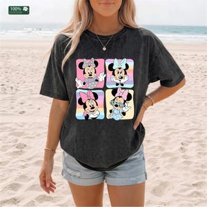 May include: A charcoal gray short-sleeve t-shirt featuring a graphic print of four distinct Minnie Mouse illustrations arranged in a square grid. Each character is set against a colorful pastel background. The fabric has a 100% cotton label.