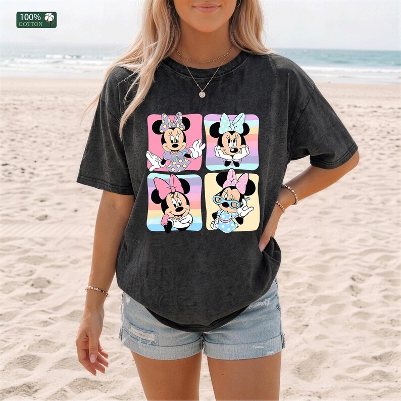 May include: A charcoal gray short-sleeve t-shirt featuring a graphic print of four distinct Minnie Mouse illustrations arranged in a square grid. Each character is set against a colorful pastel background. The fabric has a 100% cotton label.