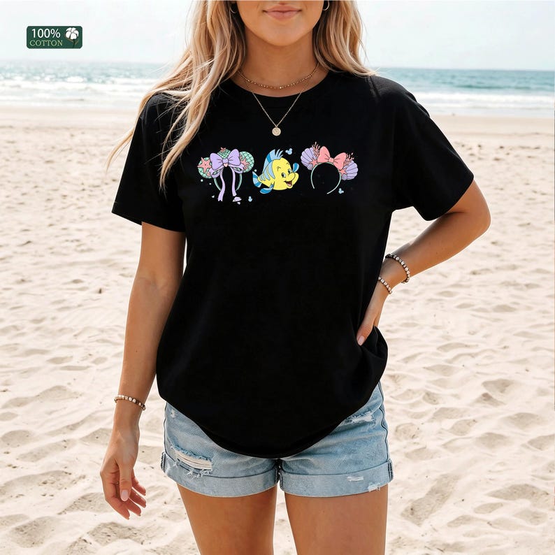 May include: A black short-sleeve t-shirt featuring a graphic print of a yellow fish character flanked by two pairs of decorative ears with seashell and bow details. The shirt includes a small green label in the corner that reads 100% COTTON.