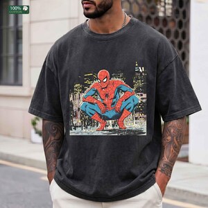 May include: A charcoal gray short-sleeve t-shirt featuring a graphic print of the comic book character Spider-Man in a crouching position against a city skyline background. The shirt has a crew neckline and a vintage-style washed fabric finish.