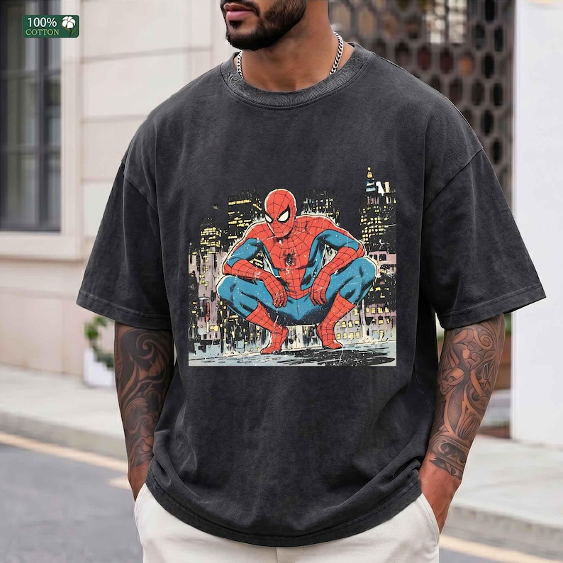 May include: A charcoal gray short-sleeve t-shirt featuring a graphic print of the comic book character Spider-Man in a crouching position against a city skyline background. The shirt has a crew neckline and a vintage-style washed fabric finish.