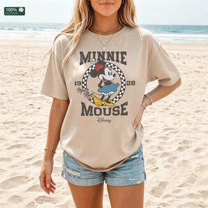 May include: A tan t-shirt featuring a graphic of Minnie Mouse. The design includes the words "MINNIE MOUSE" and "1928" in a vintage style. The shirt is made of 100% cotton and is paired with denim shorts. The background is a sandy beach.