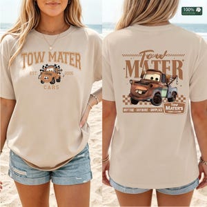 May include: A beige t-shirt featuring a graphic of the character Tow Mater from the movie Cars. The front displays a small truck illustration with the text Tow Mater Est. 2006. The back shows a larger truck graphic with the text Tow Mater.