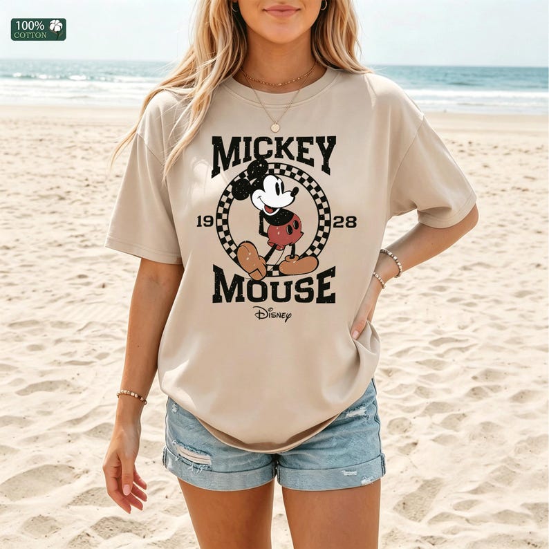 Peut inclure: Un t-shirt beige &agrave; manches courtes avec un motif de Mickey Mouse &agrave; l'int&eacute;rieur d'un cercle &agrave; damier noir et blanc. Le texte indique MICKEY MOUSE 1928 avec le logo Disney en dessous. Le t-shirt est en 100% coton avec un col rond.
