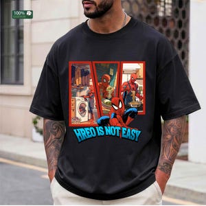 May include: A black short-sleeved t-shirt featuring a graphic print of the character Spider-Man in three separate panels performing household chores, with the text HREO IS NOT EASY printed in large blue letters at the bottom of the graphic.