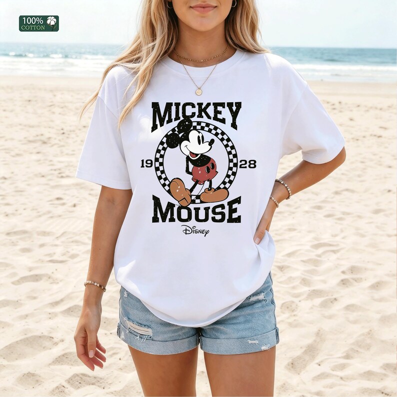 Peut inclure: Un t-shirt blanc &agrave; manches courtes avec un motif de Mickey Mouse &agrave; l'int&eacute;rieur d'un cercle &agrave; damier noir et blanc. Le texte indique MICKEY MOUSE 1928 avec le logo Disney en dessous. Le t-shirt est en 100% coton avec un col rond.