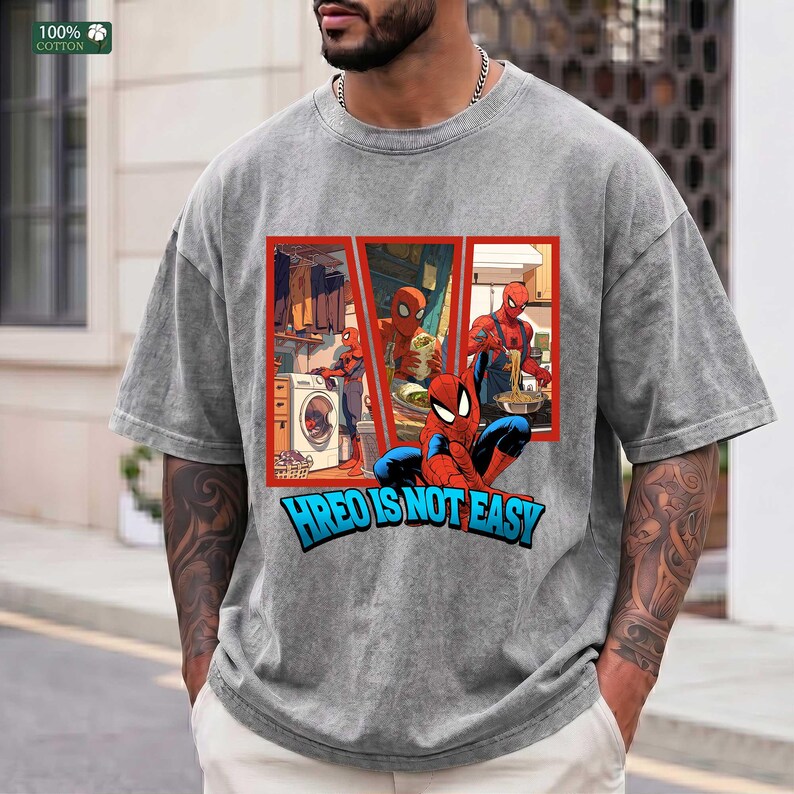 May include: A grey acid-wash t-shirt featuring a graphic print of Spider-Man performing household chores like laundry and cooking. The text below the graphic reads HREO IS NOT EASY in bold blue letters with a black outline, set against a red frame.