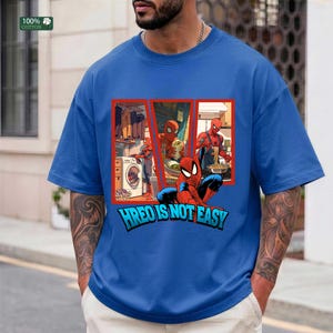 May include: A blue short-sleeve t-shirt featuring a graphic print of the character Spider-Man performing household chores like laundry and cooking. Below the graphic, the text reads HREO IS NOT EASY in bold blue letters with a black outline.