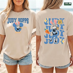 Pu&ograve; includere: Una t-shirt beige a maniche corte con una grafica di Judy Hopps sul davanti e sul retro. Il davanti mostra il testo Judy Hopps con una sagoma blu di orecchie di topo. Il retro mostra Judy Hopps, una carota, un distintivo e il testo Judy.