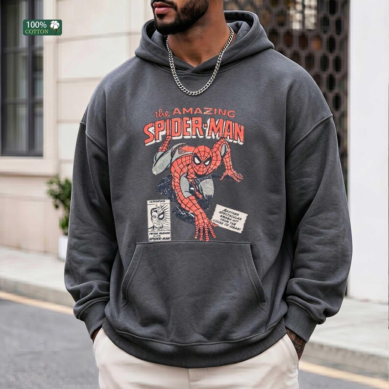 May include: A charcoal gray hooded sweatshirt featuring a vintage-style graphic of Spider-Man in a crouching pose. The text reads "the AMAZING SPIDER-MAN" with additional comic book panels and text boxes. A silver chain necklace is worn around the neck.