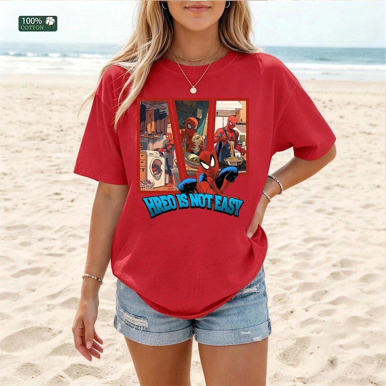 May include: A red short-sleeve t-shirt made of 100% cotton featuring a graphic print of a superhero in three panels performing daily chores, with the text "HREO IS NOT EASY" in blue letters underneath the illustration on the front of the shirt.