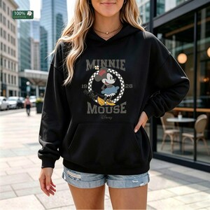 May include: Black hooded sweatshirt featuring a graphic of Minnie Mouse with the text "MINNIE MOUSE 1928". The design includes a checkered circle around Minnie and the Disney logo. The sweatshirt is made of 100% cotton.