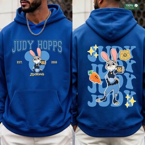Pu&ograve; includere: Una felpa con cappuccio blu reale con Judy Hopps di Zootropolis. Il davanti mostra il testo JUDY HOPPS, EST. 2016 e ZOOTOPIA con una grafica del personaggio. Il retro presenta una grande illustrazione di Judy Hopps, una carota, un distintivo e stelle.