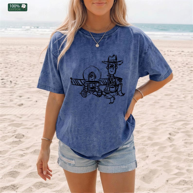 May include: A blue mineral-washed short-sleeve t-shirt featuring a black line-art graphic of two cartoon characters, one wearing a space helmet and the other a cowboy hat. The shirt has a crew neckline and a small 100% cotton label in the corner.