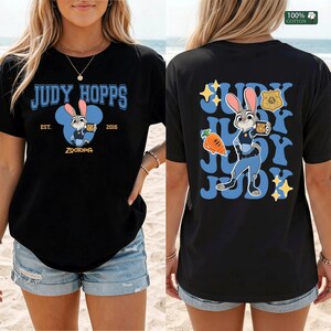 Pu&ograve; includere: Una t-shirt nera con una grafica di Judy Hopps di Zootropolis. Il davanti mostra il testo Judy Hopps, Est. 2016 e Zootopia. Il retro mostra Judy Hopps, una carota, un distintivo della polizia e il testo Judy ripetuto in carattere blu.