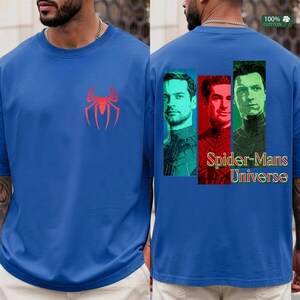 May include: A blue short-sleeve t-shirt featuring a red spider emblem on the front chest. The back displays three vertical rectangular portraits of different Spider-Man characters in teal, red, and green, with the text Spider-Mans Universe.