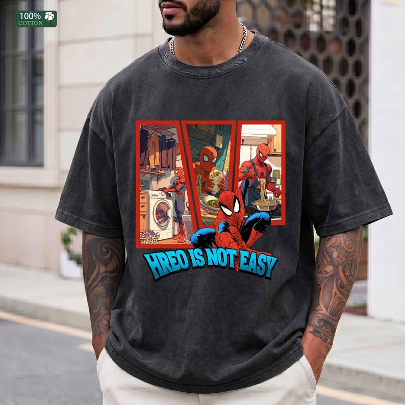 May include: A charcoal gray short-sleeve t-shirt featuring a graphic print of a red and blue superhero in three panels showing daily chores, with the text HREO IS NOT EASY printed in blue below the panels and a 100% cotton label at the neck.