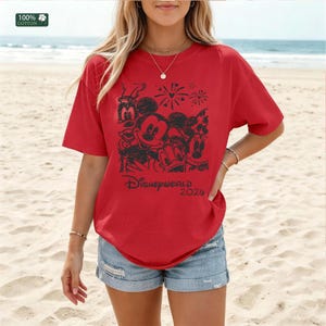 Puede incluir: Camiseta roja con un gr&aacute;fico negro de personajes de Disney, incluyendo Mickey Mouse, Goofy y Donald Duck, con fuegos artificiales y el texto "Disney World 2020." La camiseta es de 100% algod&oacute;n.