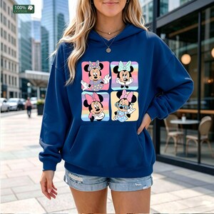 May include: A dark blue hooded sweatshirt featuring a graphic print of four squares, each containing a different illustration of Minnie Mouse with various colored bows and outfits. The upper left corner of the image displays a green 100% cotton label.