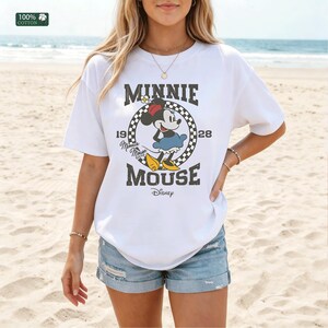 May include: White t-shirt featuring a graphic of Minnie Mouse in a black and white checkered circle, with the text "MINNIE MOUSE" and "1928 Disney". The shirt is made of 100% cotton.