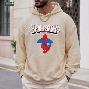 May include: Beige hoodie with a Spider-Man graphic. The design features the words "SPIDER-MAN" in a web-like frame, with Spider-Man hanging upside down. The hoodie has a kangaroo pocket and a drawstring hood.
