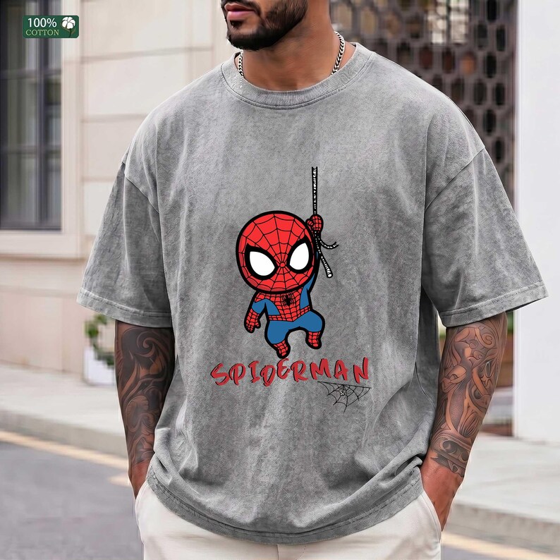 Op de afbeelding: Grijs t-shirt met een cartoon Spider-Man ontwerp. De afbeelding toont een rood en blauw Spider-Man karakter dat aan een web hangt, met het woord "SPIDERMAN" in het rood. Gemaakt van 100% katoen.
