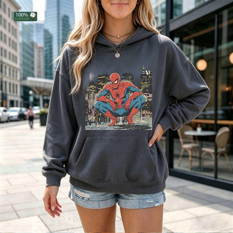 May include: A charcoal gray hooded sweatshirt featuring a graphic print of the superhero Spider-Man in a crouching position against a city skyline background. The long-sleeved pullover includes a front kangaroo pocket and a hood with drawstrings.