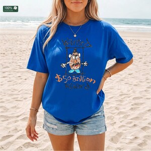 Op de afbeelding: Koning blauw t-shirt met een handgetekende afbeelding van een aardappelachtig personage met de woorden "Wanted" en "$50 Billion Reward". Het shirt is gemaakt van 100% katoen.