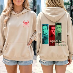 May include: A beige hooded sweatshirt featuring a red spider emblem on the front chest. The back displays three vertical rectangular portraits of men in superhero suits, colored blue, red, and green, with the text Spider-Mans Universe below.