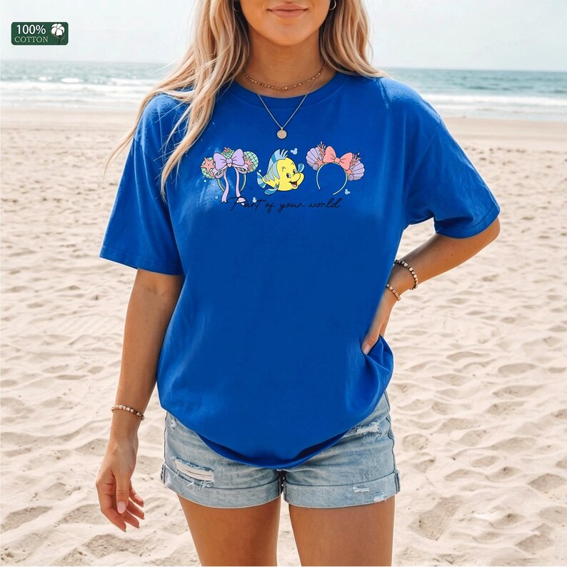May include: A royal blue short-sleeve t-shirt featuring a graphic print of two decorative headbands and a yellow fish character. The text "Part of your world" is printed below the graphic. A small label in the corner reads 100% COTTON.