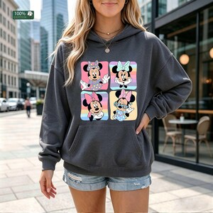 May include: A dark gray hooded sweatshirt featuring a graphic print of four squares, each displaying a different illustration of Minnie Mouse in various outfits and poses against colorful backgrounds. The garment includes a front kangaroo pocket.