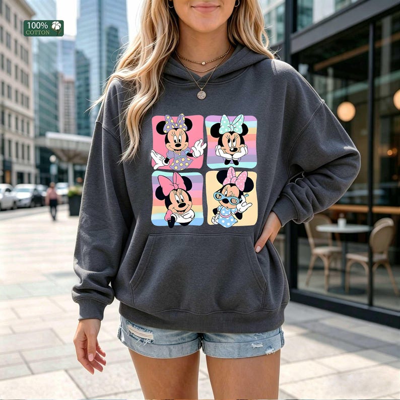 May include: A dark gray hooded sweatshirt featuring a graphic print of four squares, each displaying a different illustration of Minnie Mouse in various outfits and poses against colorful backgrounds. The garment includes a front kangaroo pocket.