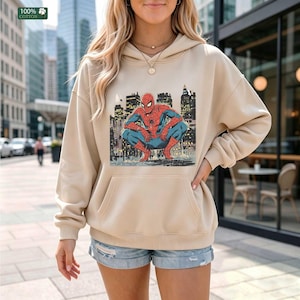 May include: A beige hooded sweatshirt featuring a graphic print of the comic book character Spider-Man crouching in front of a city skyline. The long-sleeved pullover includes a front kangaroo pocket and a hood with matching drawstrings.