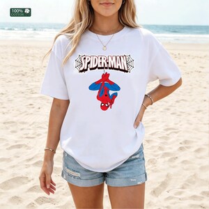 May include: White t-shirt with the text "SPIDER-MAN" in a web design, and a graphic of Spider-Man hanging upside down. The shirt is made of 100% cotton and is worn with denim shorts. The background is a beach scene.