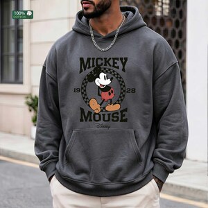 Peut inclure: Un sweat &agrave; capuche gris anthracite avec un motif de Mickey Mouse dans un cercle &agrave; damier, accompagn&eacute; du texte Mickey Mouse 1928 et Disney. Le sweat poss&egrave;de une poche kangourou et une petite &eacute;tiquette verte 100% coton dans le coin.