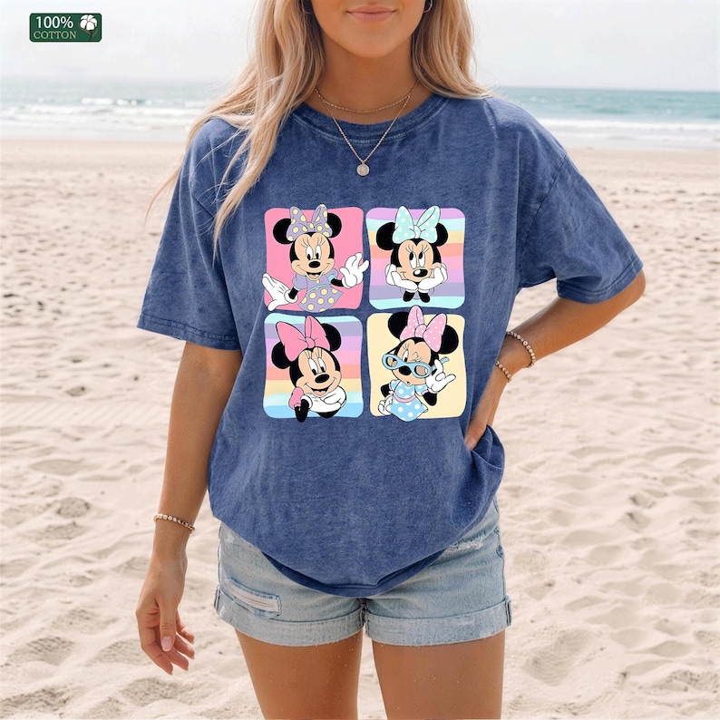 May include: A blue mineral-wash t-shirt featuring a graphic print of four squares, each containing a different illustration of Minnie Mouse wearing various bows and outfits against a pastel-colored striped background. The shirt is 100% cotton.