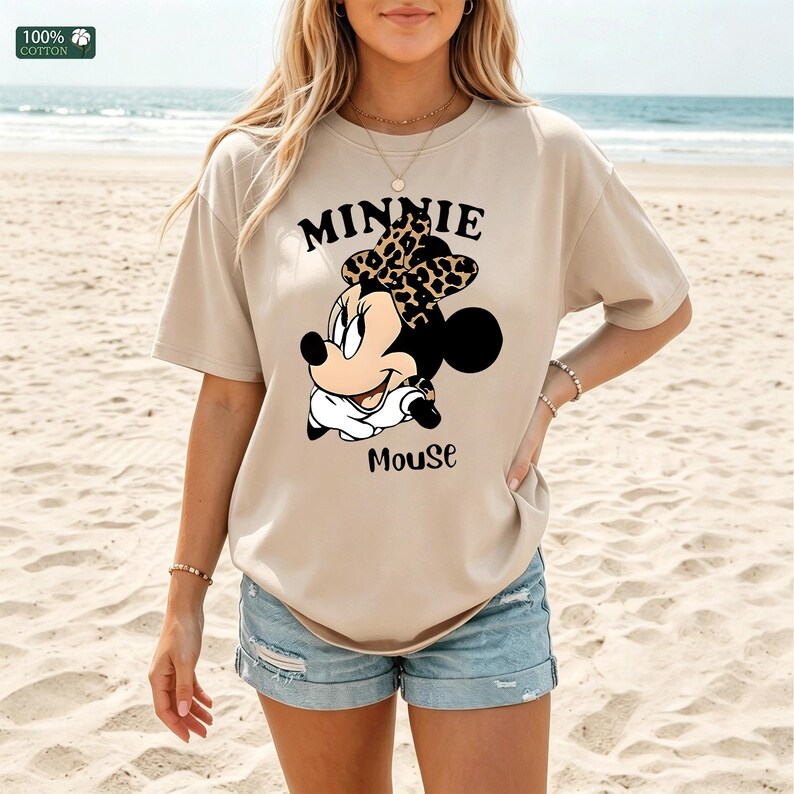 May include: Beige t-shirt featuring a Minnie Mouse graphic. The design includes the text "MINNIE" above the character's head and "Mouse" below. The bow is leopard print. The shirt is made of 100% cotton.
