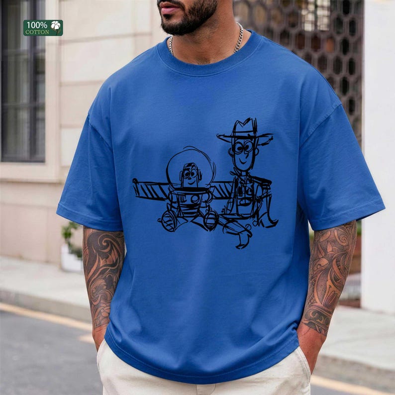 May include: A bright blue short-sleeved t-shirt featuring a black line-art graphic of a space ranger character and a cowboy character. The design is centered on the chest, showcasing a sketch-like illustration of the two iconic figures.