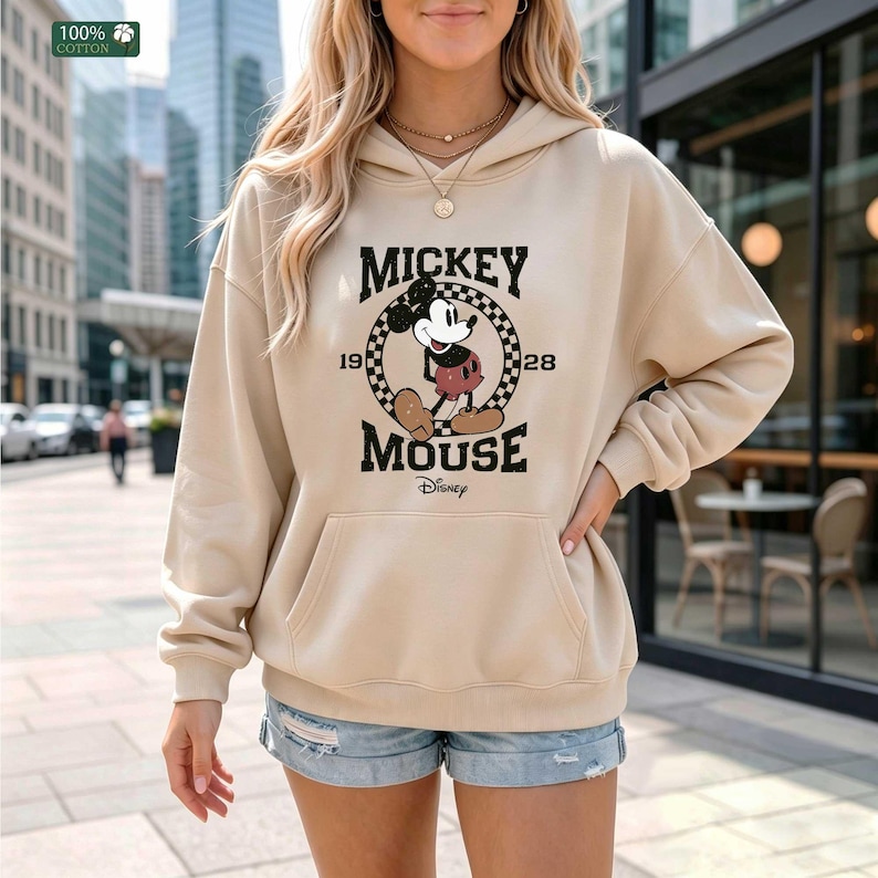 Peut inclure: Un sweat-shirt &agrave; capuche beige avec un motif de Mickey Mouse dans un cercle &agrave; damier, accompagn&eacute; du texte Mickey Mouse 1928 et Disney. Le sweat poss&egrave;de des manches longues, une poche kangourou et une &eacute;tiquette indiquant 100% coton.
