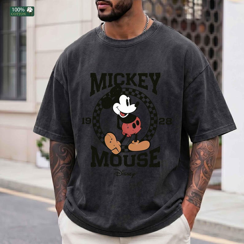 Peut inclure: Un t-shirt gris anthracite &agrave; manches courtes avec un motif de Mickey Mouse dans un cercle &agrave; damier. Le texte indique MICKEY MOUSE avec 19 et 28 sur les c&ocirc;t&eacute;s et Disney en bas. Le t-shirt pr&eacute;sente un col rond et une finition d&eacute;lav&eacute;e.