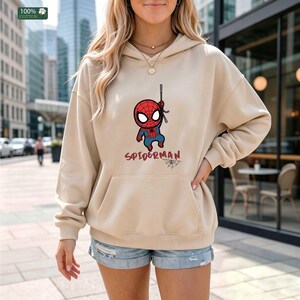 Op de afbeelding: Beige hoodie met een voorzak en een cartoon Spider-Man graphic. De graphic toont een rood-blauw Spider-Man karakter dat aan een web hangt, met het woord "SPIDERMAN" eronder. De hoodie is gemaakt van 100% katoen.