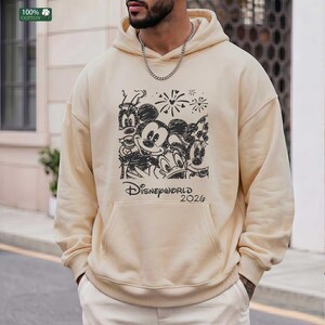 Puede incluir: Sudadera con capucha color crema con un gr&aacute;fico negro que presenta a Mickey Mouse, Donald Duck, Goofy y Minnie Mouse. El texto "Disneyworld 2026" est&aacute; debajo de los personajes. La sudadera tiene un bolsillo delantero y una capucha con cord&oacute;n.
