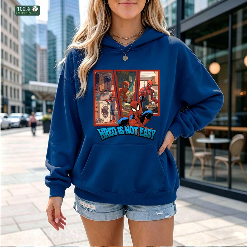 May include: A dark blue hooded sweatshirt featuring a graphic print of a superhero performing household chores like laundry and cooking. Below the graphic, the text reads "HREO IS NOT EASY" in blue and white lettering. A green "100% COTTON" tag is visible.