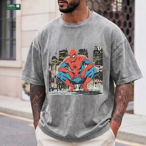 May include: A grey acid-wash t-shirt featuring a graphic print of the comic book character Spider-Man in a crouching position. The character is depicted in a red and blue suit against a dark, stylized urban cityscape background with tall buildings.