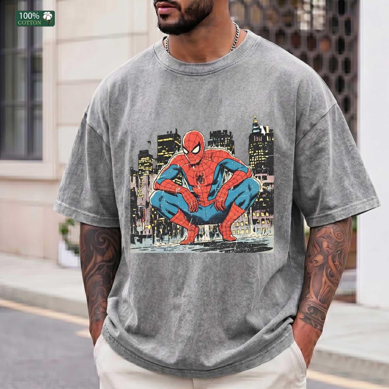 May include: A grey acid-wash t-shirt featuring a graphic print of the comic book character Spider-Man in a crouching position. The character is depicted in a red and blue suit against a dark, stylized urban cityscape background with tall buildings.
