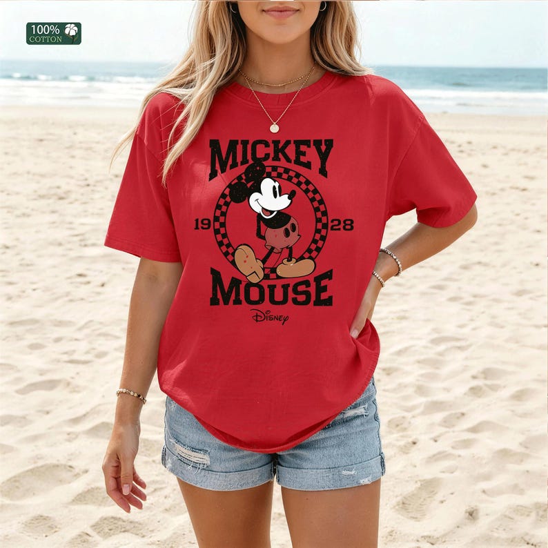 Peut inclure: Un t-shirt rouge &agrave; manches courtes avec un motif de Mickey Mouse dans un cercle &agrave; damier noir et rouge. Le texte indique MICKEY MOUSE 1928 et Disney. Une petite &eacute;tiquette dans le coin sup&eacute;rieur gauche indique une composition 100% coton.