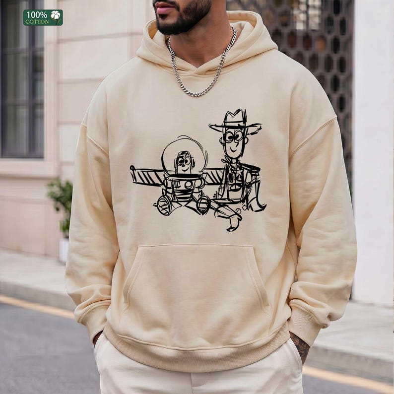 May include: A beige hooded sweatshirt featuring a black line-art graphic of two cartoon characters, a space ranger and a cowboy, printed on the front. The hoodie includes a front kangaroo pocket, long sleeves, and a drawstring hood.